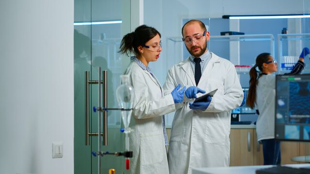 Chemist nurse explainig to doctor vaccine development in modern equipped laboratory pointing on tablet. Team of doctors examining virus evolution using high tech researching diagnosis