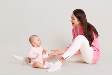 Mother playing with her daughter white sitting together on floor, kid wearing bodysuit and socks, mommy giving palm to her baby and laughing, posing isolated over white background.