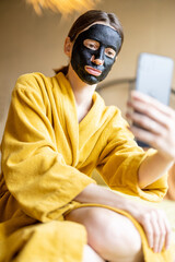 Woman in bathrobe with black cleansing mask and phone in bedroom