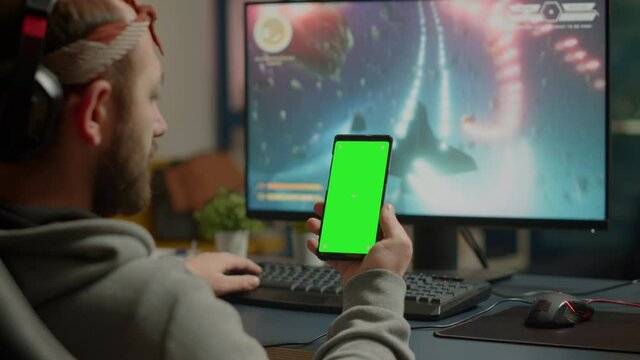 Concentrate gamer holding phone with green screen mock up chorma key display while playing games on powerful computer streaming online competition. Player using isolated desktop playing shooter games
