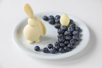 A white beautiful Easter bunny made of chocolate and an Easter egg made of white chocolate stand around blueberries on a white plate on the table for the holiday