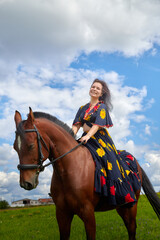 Fototapeta premium Beautiful gypsy girl on a horse in field with green glass in summer day and blue sky and white clouds background. Model in ethnic dress posing with farm animal