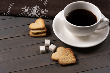 black coffee in a white cup on a white saucer with heart-shaped cookies and sugar cubes with a brown napkin with an embroidered snowflake on a brown wooden background horizontal photo side view