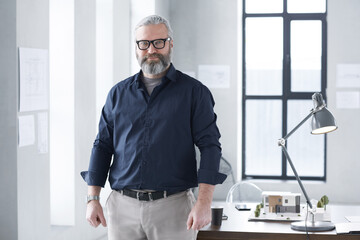 Portrait of mature bearded architect looking at camera while working at office with architectural...