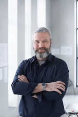 Portrait of mature bearded businessman smiling at camera while standing with his arms crossed at office