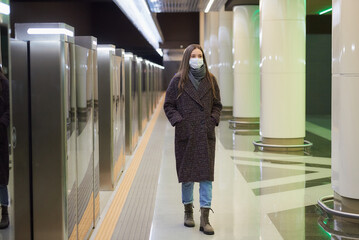 A woman in a medical face mask to avoid the spread of coronavirus is walking while waiting for a train on the subway platform. Girl in a surgical mask is keeping social distance.