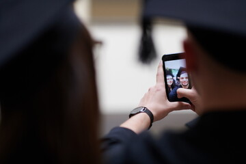 group of happy international students in mortar boards and bachelor gowns with diplomas taking selfie by smartphone