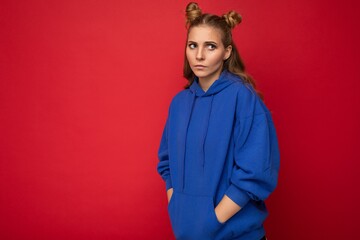 Portrait of young sad upset attractive winsome blonde woman with two horns with sincere emotions wearing casual bright blue hoodie isolated on red background with free space