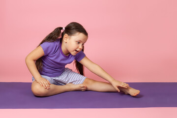 Fototapeta na wymiar full-length portrait of a preschool child doing gymnastics and doing side bends while sitting on a purple sports mat, isolated on a pink background.