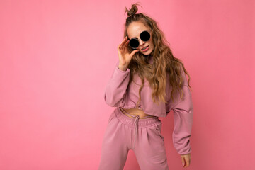 Photo shot of beautiful young dark blonde woman wearing casual clothes and stylish sunglasses isolated over colorful background looking at camera