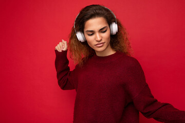 Fototapeta premium Photo of beautiful happy smiling young brunette curly woman wearing dark red sweater isolated over red background wall wearing white bluetooth headphones listening to music and having fun looking to
