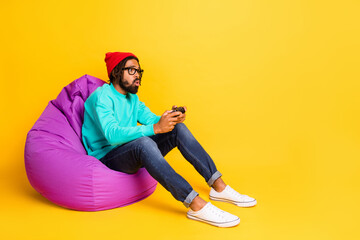 Full size profile photo of brunette optimistic guy playstation wear cap spectacles pullover jeans isolated on yellow background © deagreez