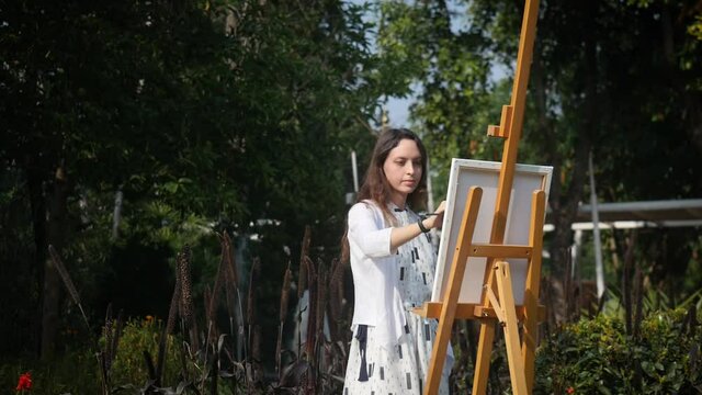 Girl artist in a white dress does oil painting on canvas in the flower garden. With loose hair, concentrated, focused, hard-working and professional.