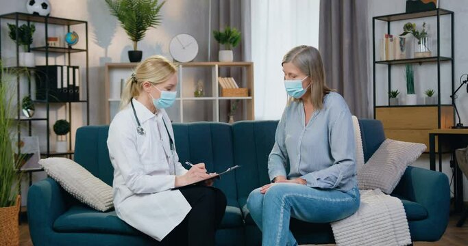 Healthcare concept where good-looking experienced female medical worker in facial mask and white gawn visiting female patient at home during desease