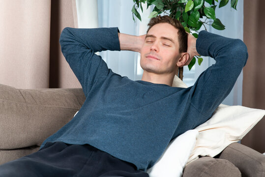 Portrait of relaxed happy guy, young handsome sleeping, enjoying, resting man having a nap at comfortable couch at home with his hands behind head and closed eyes smiling, relaxing at weekend. Freedom