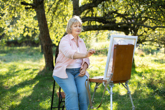 Creativity, hobby, art, leisure and people concept. Portrait of happy senior woman artist with paint brush, drawing on easel outdoors in the open air in green park in summer.