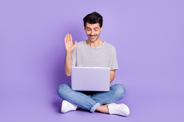 Photo of guy sit floor hold netbook look screen wave hand wear white t-shirt jeans footwear isolated purple background