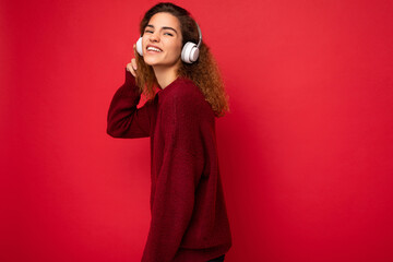 Fototapeta premium Beautiful happy smiling young brunette curly woman wearing dark red sweater isolated over red background wall wearing white bluetooth headphones listening to music and having fun looking at camera