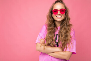 Photo shot of beautiful smiling happy young dark blonde woman wearing casual clothes and stylish sunglasses isolated over colorful background looking at camera