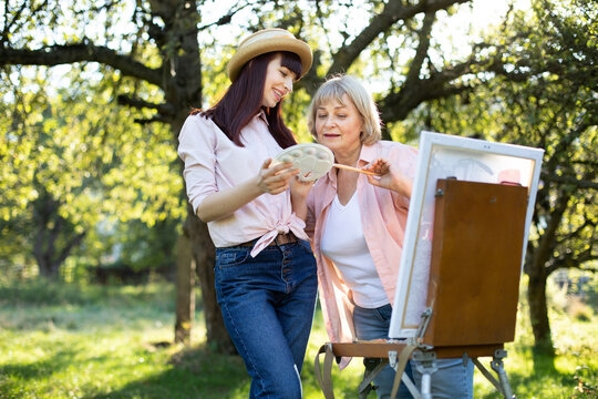 Close up outdoor portrait of senior 60-aged woman and her young daughter, painting together using easel on sunny day in the garden. Elderly mother and young daughter choosing a paint on a palette.