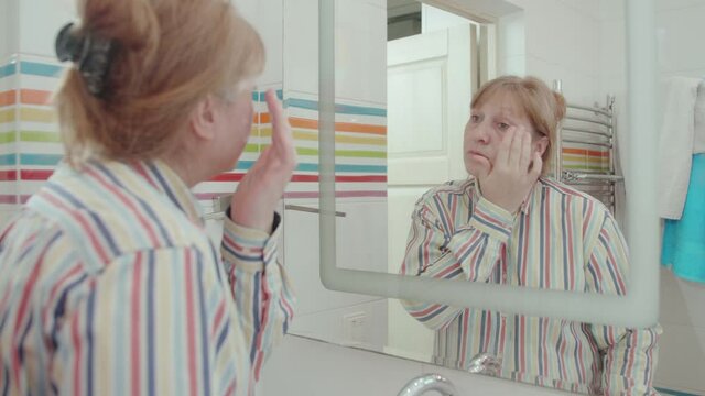 Portrait senior woman applying moisturizer to her face looking in the mirror. Wonderful senior model with blonde hair in white shirt in white bathroom.