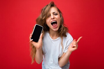 Photo of beautiful joyful smiling young blonde woman good looking wearing white t-shirt standing isolated on red background with copy space holding phone showing smartphone in hand with empty screen - obrazy, fototapety, plakaty