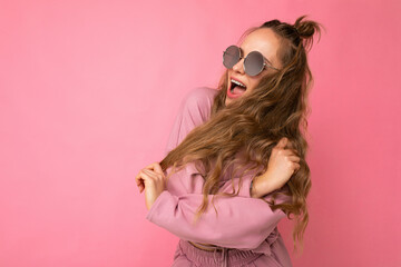 Shot of beautiful happy emotionsl young dark blonde curly woman isolated over pink background wall wearing casual pink sport clothes and stylish sunglasses looking to the side and having fun
