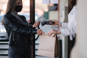 Young woman standing outdoors in mask and paying bill through smartphone using NFC technology in a...