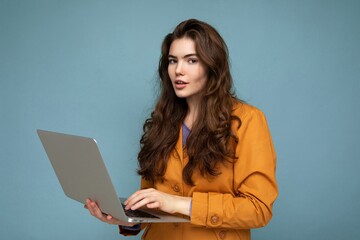 Close-up portrait of Beautiful brunet curly young woman holding netbook computer looking at camera wearing yellow jacket typing on keyboard isolated on blue background