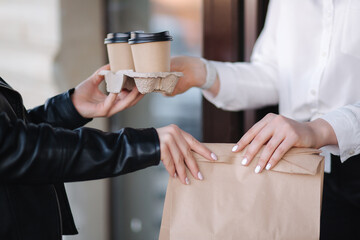 Customer in face mask stand outdoors and take package with coffee and food from cashier in mask at cafe. Take away food. Close up of hands. Quarantine