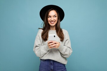 Beautiful young brunette woman thinking wearing black hat and grey sweater holding smartphone looking to the side texting isolated on background