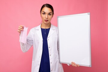 Female person doctor in a white medical coat holding blank board with copy space for text and protective mask isolated on pink background. Epidemic concept