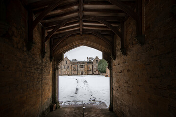 Looking through ancient gatehouse to view of abandoned old derelict run down eerie Manor House boarded up in dark creepy winter snow scene. Archway leading to deserted mansion.