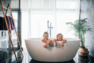 Two kids having fun and washing themselves in the bath at home. Posing for a camera