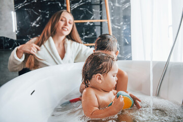 Having fun. Young mother helps her son and daughter. Two kids washing in the bath