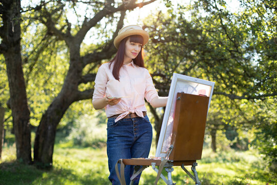 A young woman artist holds a brush and palette and paints a picture on an easel in the rays of the sunset outside in green summer garden. The female painter drawing a picture in a park.