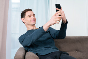 Happy handsome guy, young man blogger is sitting on bed at home in bedroom and taking selfie, picture of himself for social media on his cell mobile phone, looking at frontal camera of smartphone. 