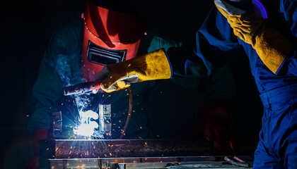 a factory worker wearing a green mechanic coveralls and safety helmet welding metalwork at night time in a factory while his colleague looking at a spark from welding