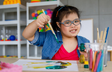 Young Asian down syndrome little girl sitting at desk with colors and painting with concentrated...