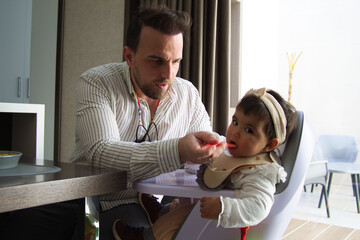 homosexual father feeding the porridge to the little daughter sitting in his chair.