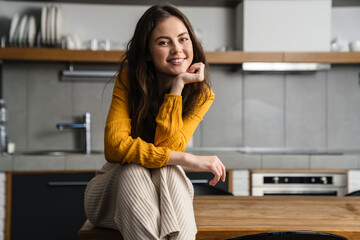 Young brunette woman smiling at camera while sitting on table in kitchen