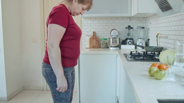 Senior woman in red t-shirt standing on weight scale and rejoicing by weight loss being in white kitchen interior. 