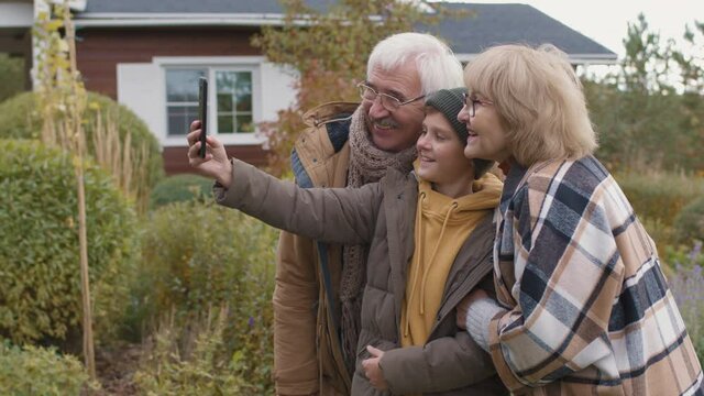 Medium shot of senior Caucasian woman, man and their teenage grandson wearing warm casual clothes standing in garden near house and making selfie using telephone camera