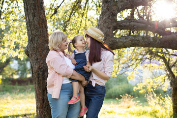 Fototapeta premium Multi generation family in the garden in summer. Retired grandmother holds beautiful little girl on hands, while she is plaing with young pretty mom, touching noses.