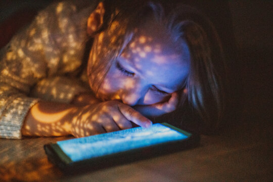 Little Girl Lying Under The Bed And Looking At Something On The Mobile