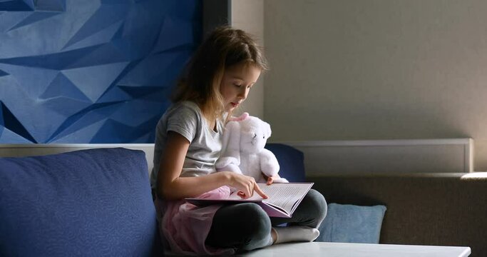 Cute little girl eading a book with stuffed teddy bunny toy and smiling sitting on the sofa in the living room. Stay at home during coronavirus covid-19 pandemic