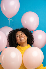 Happy pleased African woman with curly hair holding a lot of pink balloons, enjoys cool party, wears yellow sweater, celebrates birthday, looking up standing over studio blue background. Festive event