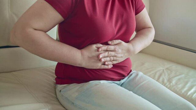 Portrait of a caucasian woman with blonde hair sitting on sofa having strong stomach ache hands on abdomen