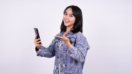 Asian women smiling wearing jeans jacket and holding and pointed at the phone with isolated white background