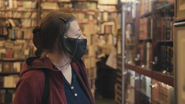 Serious woman in glasses and mask against virus chooses painting to buy in an antique store. Concept of collectibles, hobbies in art during epidemics and pandemics of virus